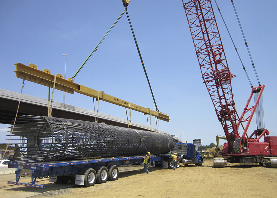 A crane lifts a round prefabricated rebar cage from a flatbed truck on a construction site.