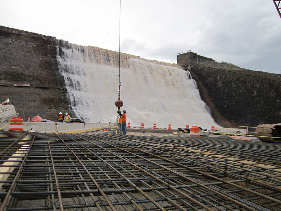 A old dam spillway sheds water normally in the background while workers prepare a new concrete spillway next to it.