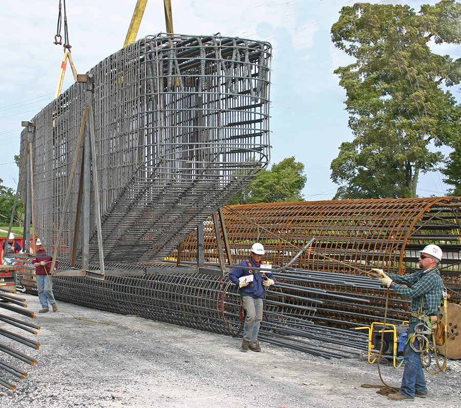 A crane moves a rebar bridge pier cage in a construction staging area.